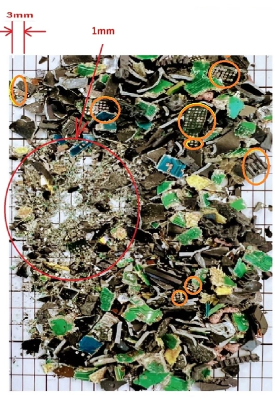 電子回路基板などが細かくシュレッダー処理された廃棄物の山が写っています。様々な色の破片の中に、小さな電子部品が見られ、赤い円と矢印で特定の箇所が強調されています。グリッド上でサイズが示されています。