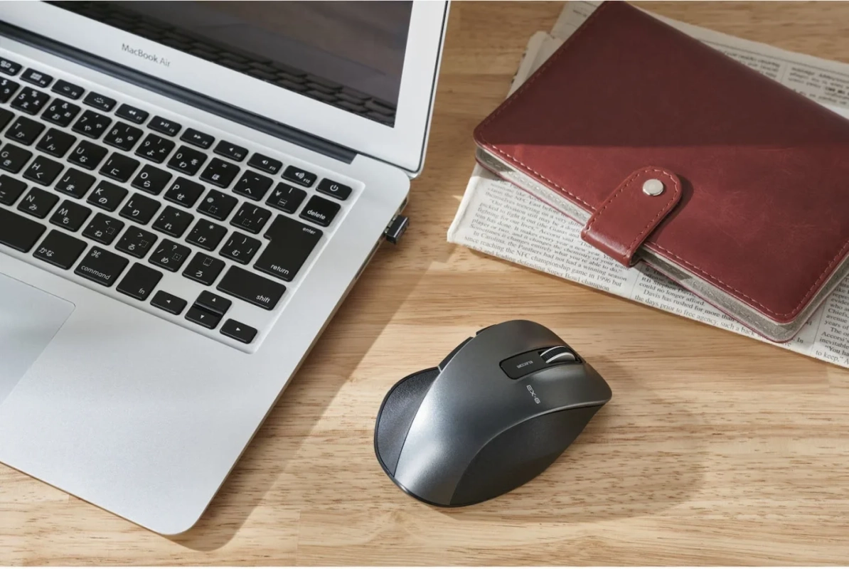 MacBook AirとELECOMのワイヤレスマウス、そして革製の手帳が木目のデスクに置かれたビジネスシーンの画像です。