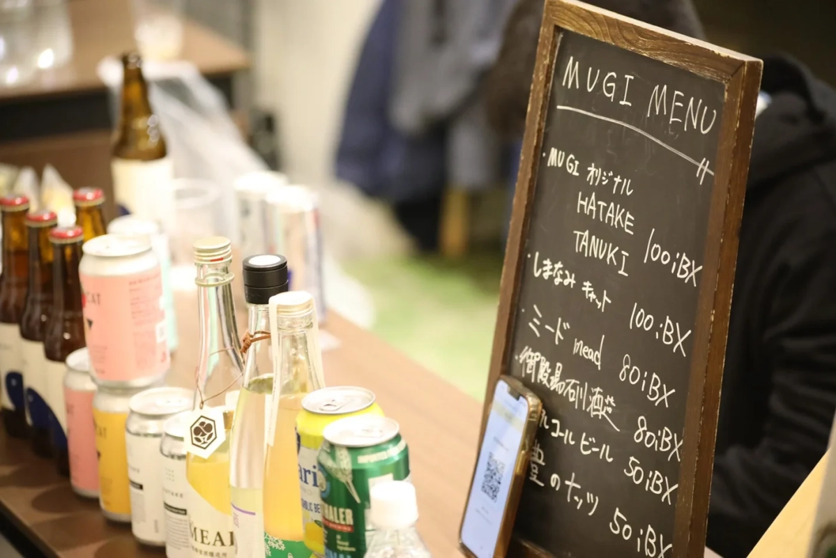 カウンターに並べられた様々なボトルや缶の飲み物と、手書きのMUGIメニューが書かれた黒板が写っています
