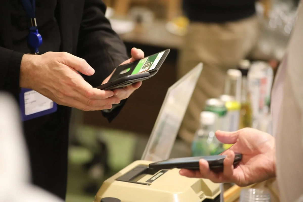 会場のような場所で、2人の人物がスマートフォンやカードを使って何らかの登録や支払いのやり取りをしている様子