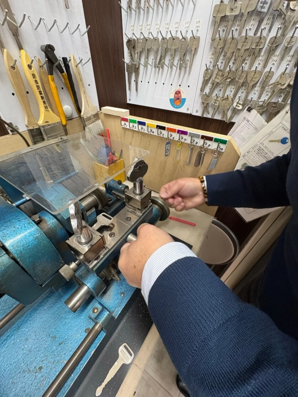 青い合鍵作成機で鍵を複製する作業風景。壁には多数の鍵が番号別に整理されている。