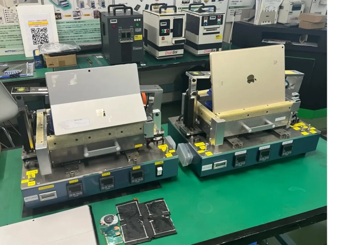 電子機器のデータ消去やリサイクルが行われている作業場の様子。Microsoft SurfaceとiPadが専用装置にセットされ、背景にはデータ破壊装置「CrushBox」やGIGAスクール構想関連の掲示が見える。