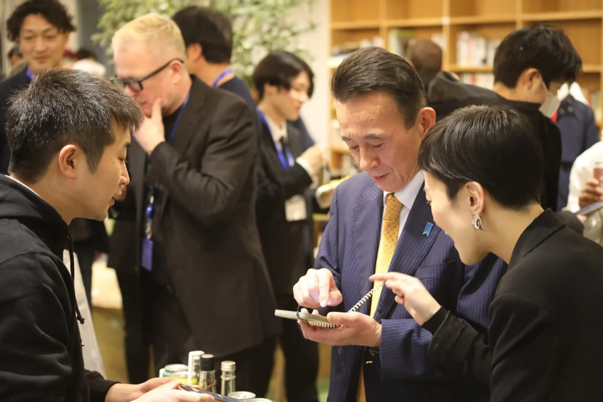 屋内のイベントで、スーツ姿の男性がスマートフォンを操作し、隣の女性が画面を指差して説明している様子