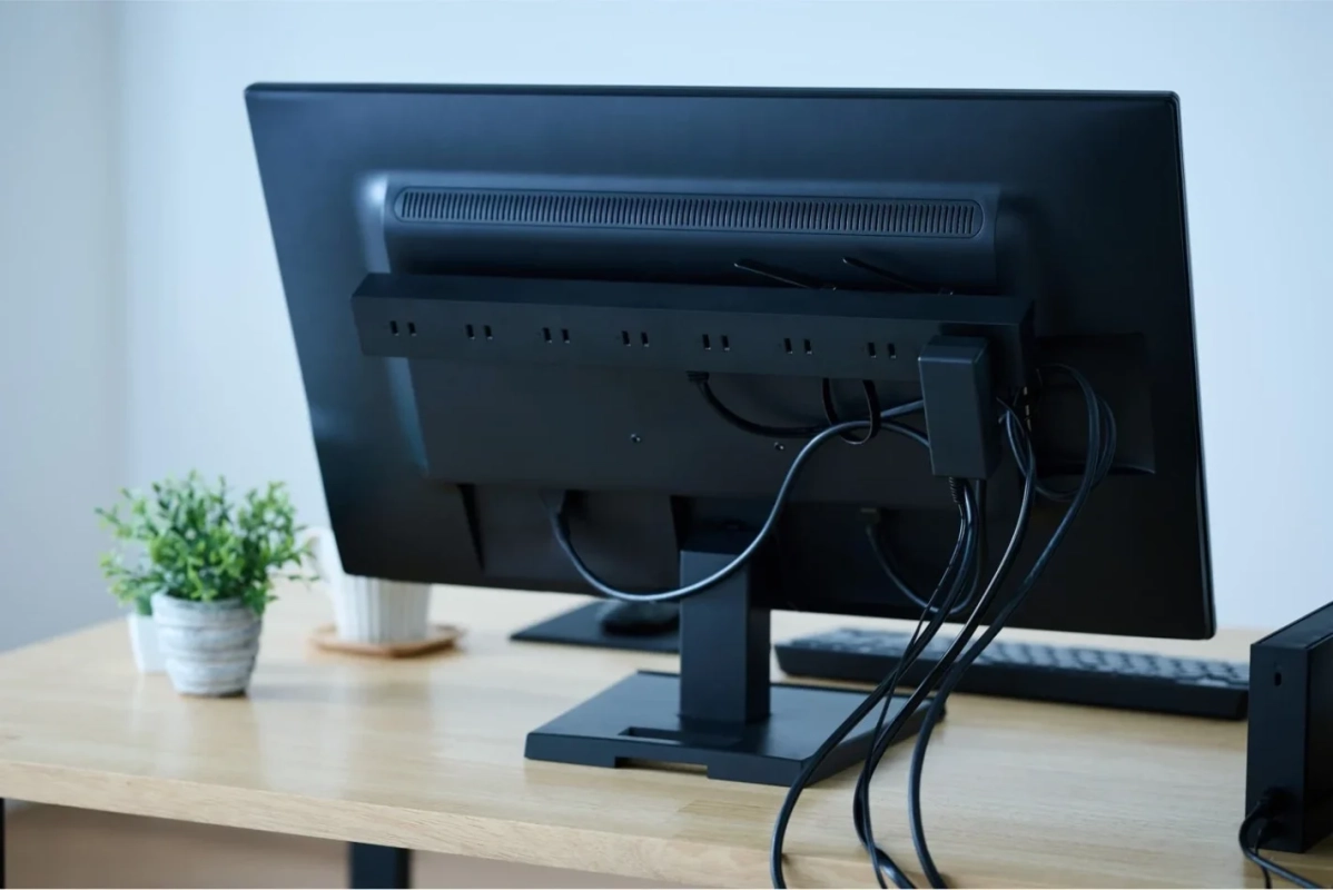 木製のデスクに置かれたパソコンモニターの背面が写っており、多くのケーブルが整理されて接続されています。