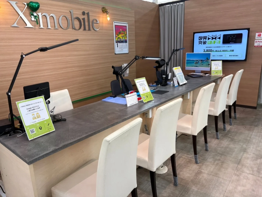 Xmobile店舗のカウンターエリア。複数の顧客席とスタッフ用デスクが並び、大型モニターにはWi-Fiの料金プランが表示されている。