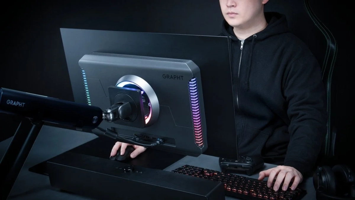 黒いパーカーを着た男性が、RGBライティングが特徴的なゲーミングモニターと周辺機器が並ぶデスクでPCを操作しています。