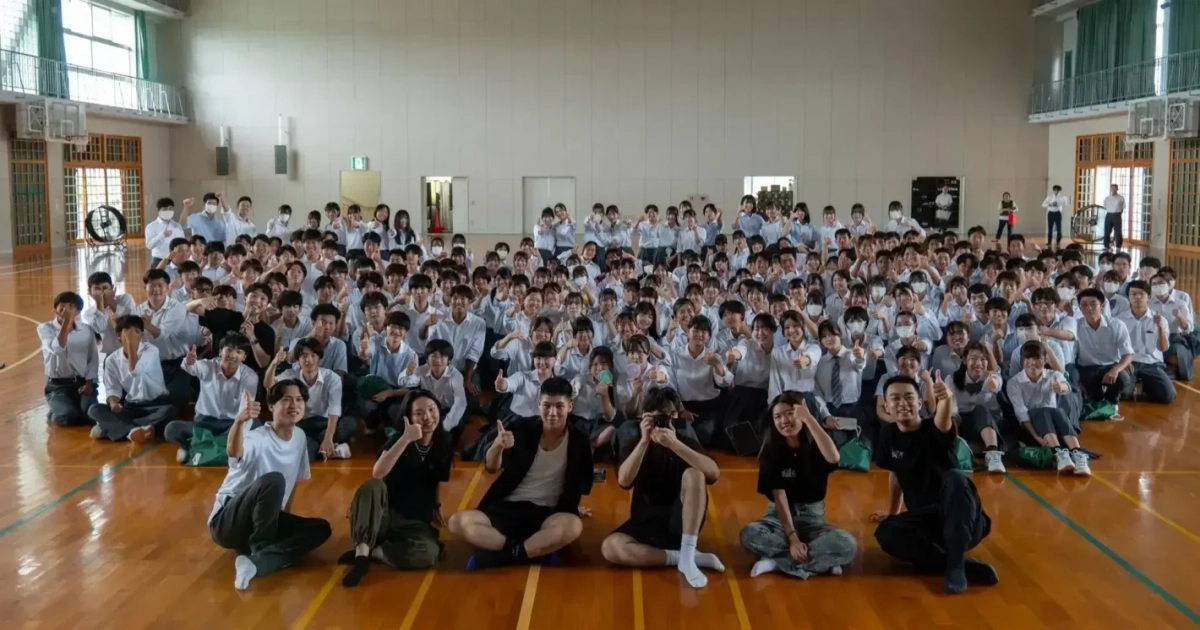 体育館で多くの学生が笑顔でサムズアップポーズをとっている集合写真