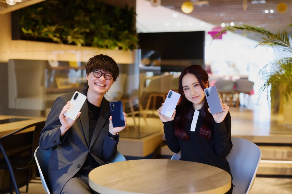 カフェのようなモダンな空間で、笑顔の男女が複数色のスマートフォンを手に持ち、カメラにアピールしています。新製品の紹介やプロモーションに見えます。