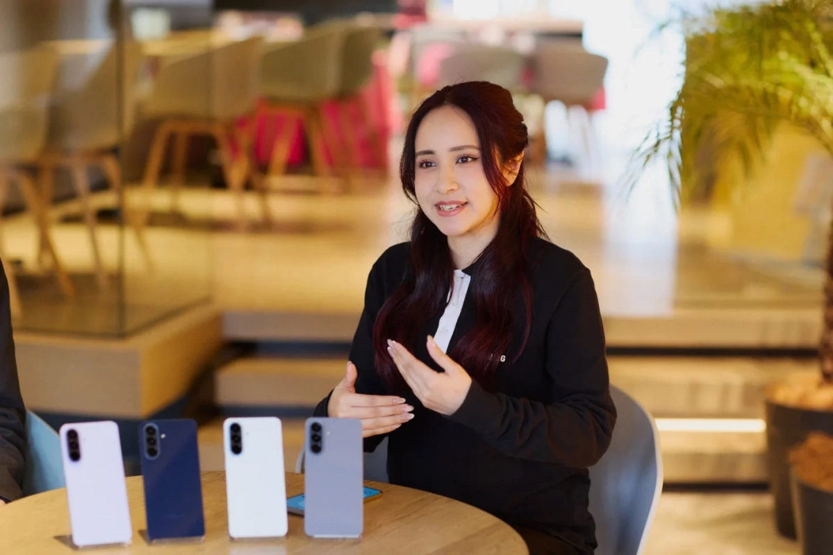 屋内の会場で、一人の女性がスマートフォン製品について説明している様子です。テーブルには複数の色のスマートフォンが展示されており、女性は身振り手振りを交えて話しています。