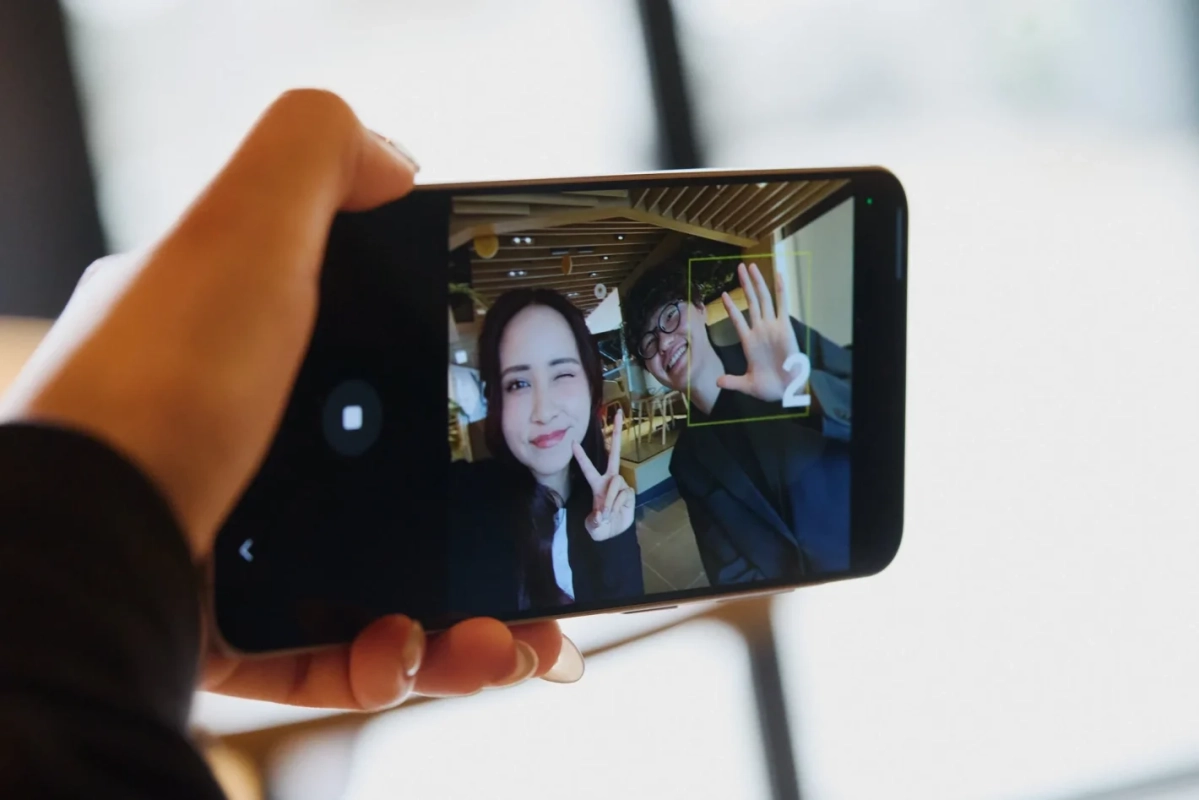 スマートフォンで自撮りをする男女の様子が映し出されています。女性はウィンクとピースサイン、男性は笑顔で手を広げ、画面には数字の「2」が表示されており、撮影の瞬間の楽しげな雰囲気が伝わります。