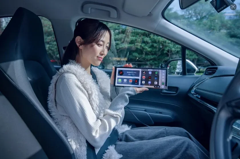 車の助手席に座る若い女性が、様々なアプリが表示された大型のポータブルスクリーンデバイスを手に持っています