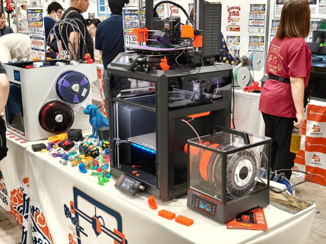 日本ホビーショーの会場で、複数の3Dプリンターが展示され、カラフルな3Dプリント作品が多数並べられている様子