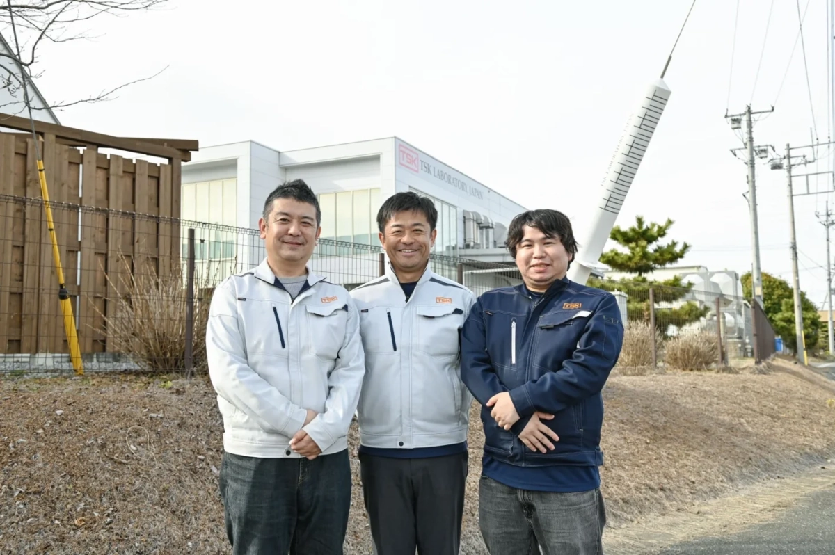 TSK LABORATORY JAPANの建物前で、作業着姿の男性3人が笑顔で立っています。背景には巨大な注射器のモニュメントも見え、研究施設であることが伺えます。