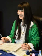 緑色のカーディガンを着た長い黒髪の女性が、書類を前にして笑顔を見せています。ペンを持ち、楽しげな表情で作業に集中している様子が伺える一枚です。