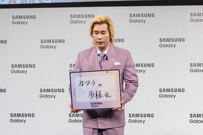 金髪の男性がピンクのスーツ姿で、「カタチの多様化」と書かれたタブレットを掲げている