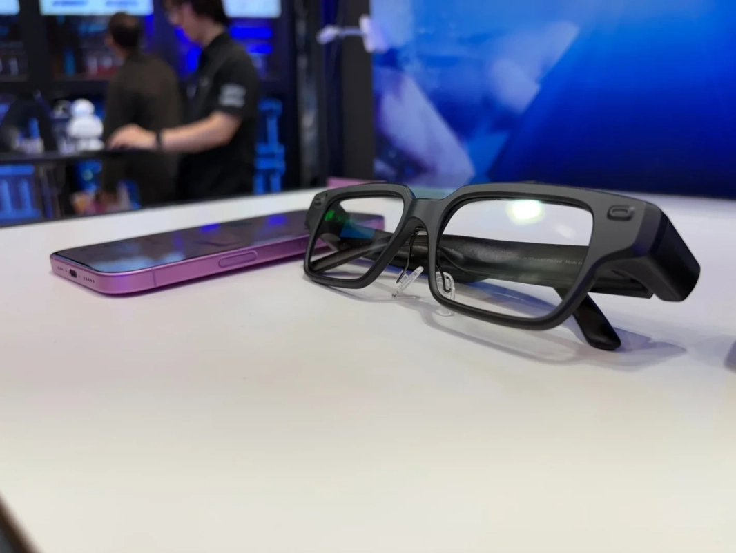 白いテーブルに黒いスマートグラスと紫色のスマートフォンが置かれています。背景にはぼやけた人々と青いディスプレイが見え、テクノロジーイベントの雰囲気です。