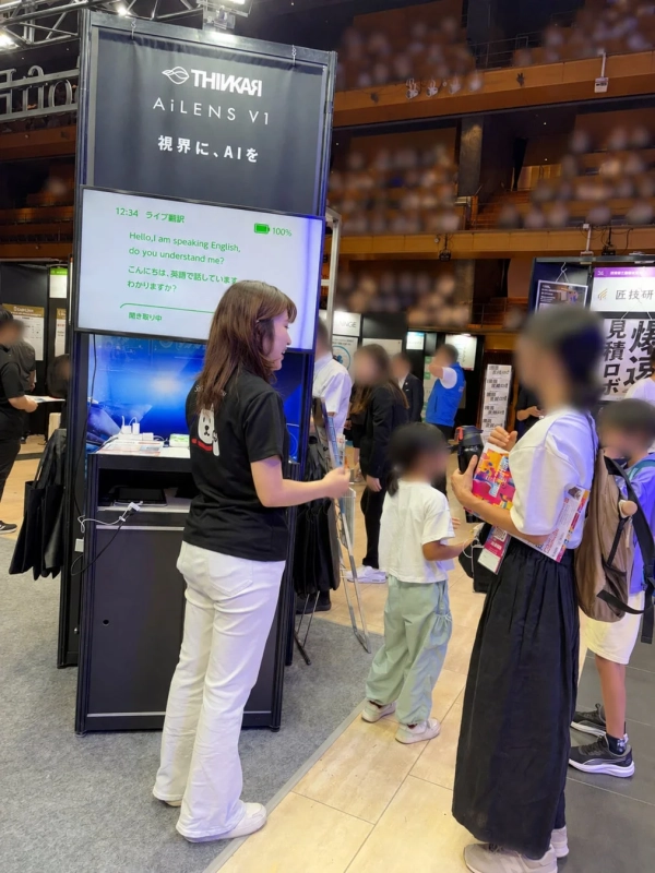 展示会ブースで、AIライブ翻訳システム「AILENS V1」のデモンストレーションが行われている様子。女性スタッフが説明しており、画面には英語と日本語の翻訳が表示されている。来場者が興味深そうに見学している。