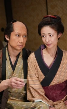 和服を着た男女が室内で並んで座っている画像です。男性は髷のような髪型で小さな杯を持ち、女性は伝統的な髪型で落ち着いた表情をしています。時代劇の一場面のような雰囲気です。