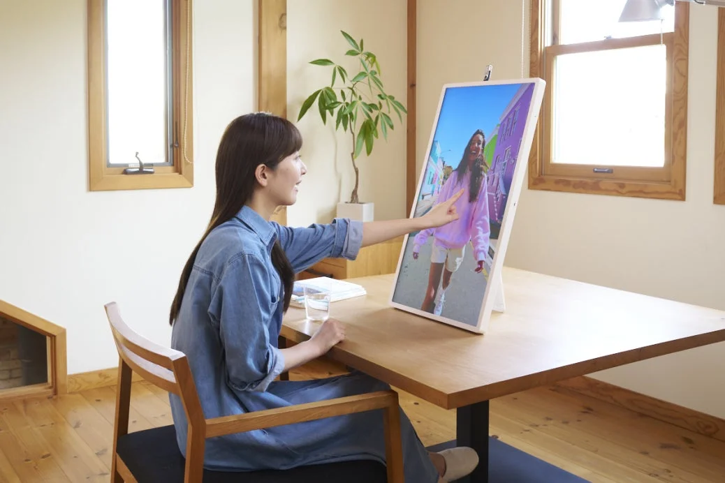 明るい部屋の木製デスクで、一人の女性が大型の縦型スマートディスプレイを操作しています。画面には屋外で笑顔を見せる別の女性が映し出されており、指を差してインタラクションしている様子です。