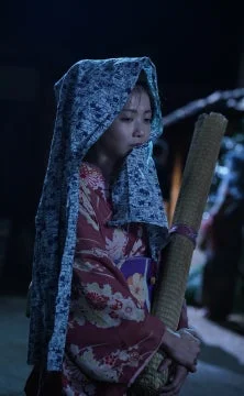 和服を身につけ、頭巾をかぶった女性が、暗闇の中で大きな巻き物のようなものを持って立っています。表情はやや沈痛で、神秘的な雰囲気を醸し出しています。